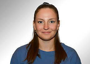 Frontales Porträt einer Person mit langen, braunen, zurückgebundenen Haaren; dezentes Lächeln. Trägt blaues T-Shirt und mehrere Ohrringe. Heller, neutraler Hintergrund.