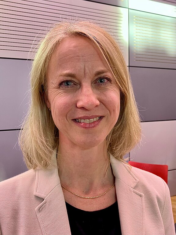 Porträtfoto einer lächelnden, blonden Frau mit schulterlangem Haar, hellem Blazer und schwarzem Oberteil; sie blickt in die Kamera vor neutralem Innenraumhintergrund.