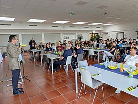 Eine Person steht am Mikrofon und hält eine Rede vor einem Publikum in einem großen Raum. Die Zuhörer, die an Tischen sitzen, hören aufmerksam zu. Auf den Tischen sind Getränke und Snacks platziert.