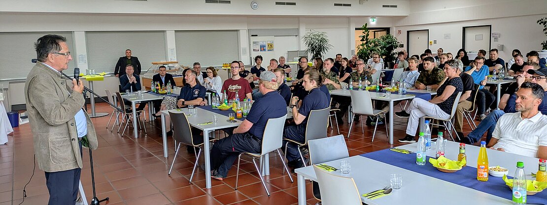 Eine Person steht am Mikrofon und hält eine Rede vor einem Publikum in einem großen Raum. Die Zuhörer, die an Tischen sitzen, hören aufmerksam zu. Auf den Tischen sind Getränke und Snacks platziert.