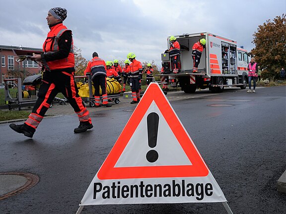 Auf dem Bild ist ein Warnschild mit der Aufschrift "Patientenablage" zu sehen. Im Hintergrund arbeiten mehrere Rettungskräfte in orangefarbener Kleidung, während ein Rettungswagen bereitsteht. Die Szene vermittelt eine Notfallsituation, in der Patienten angemeldet werden.