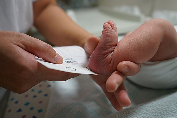 Eine Hand hält ein Dokument, während sie einen kleinen Fuß eines Neugeborenen berührt. Der Fuß ist leicht gebogen und zeigt die kleinen Zehen. Die Szene vermittelt einen medizinischen Kontext, möglicherweise einen Identifikationsprozess bei einem Baby.