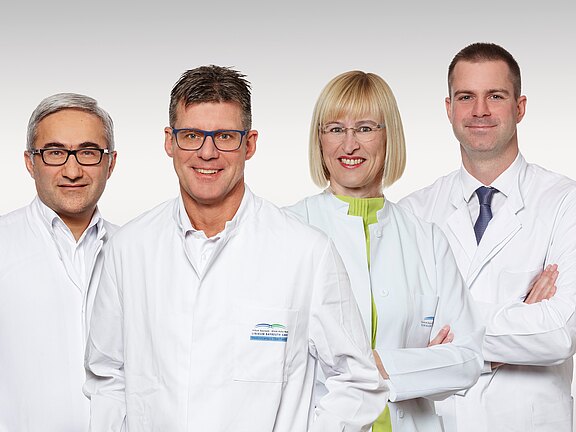 Vier Ärztinnen und Ärzte in weißen Kitteln posieren nebeneinander vor neutralem Hintergrund und lächeln in die Kamera; zwei tragen Brillen, einer eine Krawatte. Teamfoto einer medizinischen Praxis.