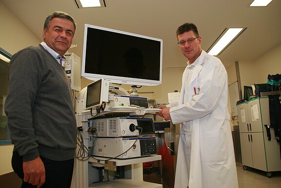 Zwei Männer in einem Klinikraum stehen an einem medizinischen Gerätenwagen mit großem Monitor. Der Mann im weißen Kittel bedient die Endoskopie- und Diagnosegeräte, offenbar zur Vorbereitung oder Demonstration medizinischer Untersuchungen.