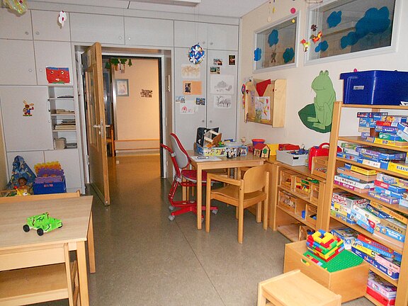 Kinderspielraum mit kleinen Tischen und Stühlen, Regalen voller Brettspiele und Bauklötze, Basteltisch mit Material; heller, ordentlicher Raum zum Spielen und Basteln.