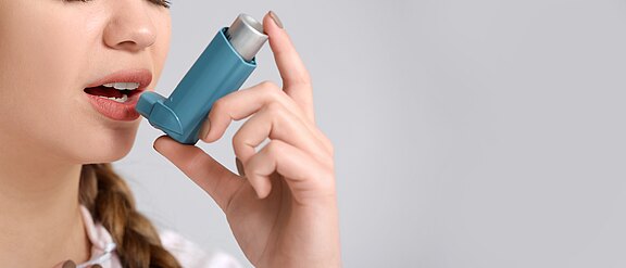Nahaufnahme: Eine Person hält einen blauen Asthma-Inhalator an den leicht geöffneten Mund und setzt zum Sprühstoß an. Der Hintergrund ist neutral und unscharf.