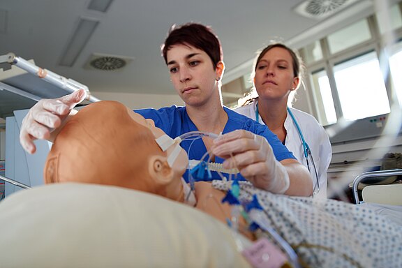Zwei Fachkräfte in Krankenhausuniformen kümmern sich um eine Trainingspuppe. Eine Person platziert einen Schlauch an der Puppe, während die andere aufmerksam zuschaut. Die Szene zeigt eine medizinische Ausbildung oder Simulation zur Patientenversorgung.