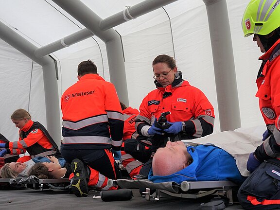 Im Bild sind Sanitäter in leuchtenden Overalls zu sehen, die sich um mehrere verletzte Person kümmern. Die Sanitäter arbeiten in einem Zelt und nutzen medizinische Geräte. Einige der Verletzten liegen auf Tragen und erhalten erste Hilfe.
