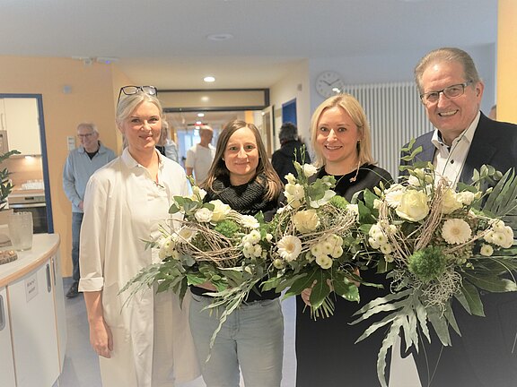 Vier Erwachsene posieren in einem Krankenhausflur. Zwei Frauen in der Mitte halten große Blumensträuße mit weißen Blüten. Links steht eine Person im weißen Kittel, rechts ein Mann im Anzug. Die Gruppe lächelt; die Szene wirkt wie eine Gratulation oder Ehrung.