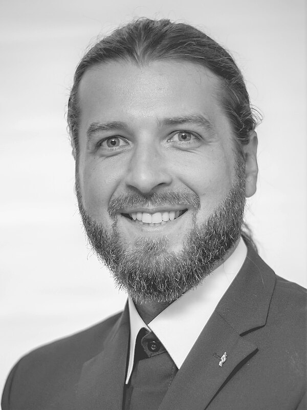 Schwarzweiß-Porträt eines lächelnden Mannes mit Vollbart und zurückgebundenen langen Haaren. Er trägt Anzug mit Hemd und Sakko, eine kleine Anstecknadel am Revers ist sichtbar. Kopf und Schultern frontal, neutraler Hintergrund.