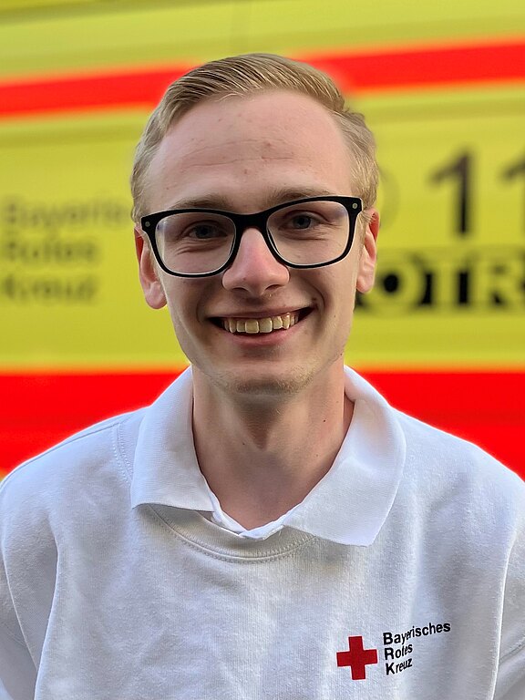 Porträt eines lächelnden jungen Mannes mit Brille in weißem Polohemd mit Aufschrift „Bayerisches Rotes Kreuz“, vor einem gelb-roten Rettungsfahrzeug.