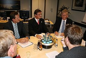 Eine Gruppe von fünf Männern sitzt an einem Tisch in einem Konferenzraum. Sie diskutieren und haben Notizen vor sich. In der Mitte des Tisches steht ein Behälter mit Wasserflaschen und Gläsern. Der Raum wirkt professionell und gut organisiert.