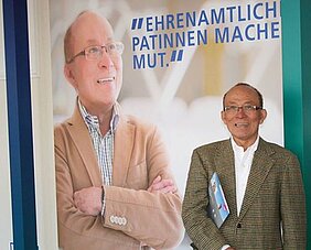 Ein lächelnder Mann steht neben einem großen Plakat, auf dem er ebenfalls abgebildet ist. Der Hintergrund des Plakats ist schlicht und trägt den Text „Ehrenamtlich Patinnen machen Mut.“ Der Mann trägt eine Brille und hält eine Broschüre in der Hand.