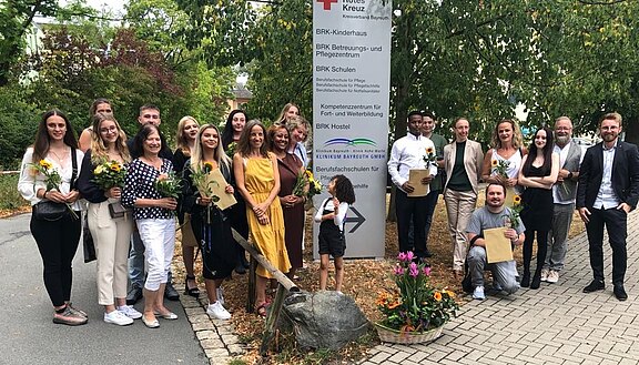 Eine große Gruppe junger Erwachsener posiert vor einem BRK-Schild im Freien. Viele halten Sonnenblumen und Umschläge. Es wirkt wie eine Abschlussfeier oder Zeugnisvergabe einer Pflegeausbildung. Alle lächeln in die Kamera.