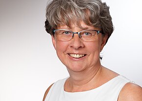 Porträt einer lächelnden Frau mit kurzen, grauen Haaren, die eine brille trägt. Sie hat einen freundlichen Ausdruck und trägt ein ärmelloses, helles Oberteil. Der Hintergrund ist neutral und hell.