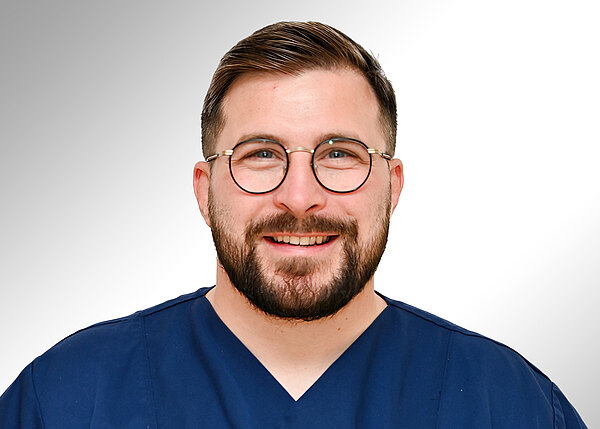 Porträt eines lächelnden Mannes mit runder Brille und Bart, in dunkelblauer medizinischer Kleidung, vor hellem neutralem Hintergrund.