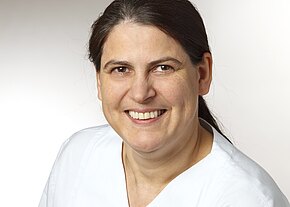 Porträt einer lächelnden Frau mit dunklen, zurückgebundenen Haaren in weißer Arbeitskleidung vor neutralem Hintergrund; Kopf- und Schulteransicht.