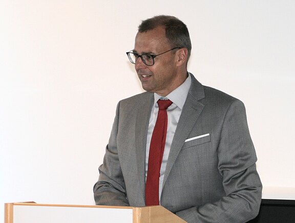 Ein Mann im grauen Anzug mit roter Krawatte spricht am Rednerpult. Er trägt eine Brille; neutraler, heller Hintergrund.