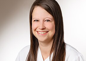 Porträt einer lächelnden jungen Frau mit langem braunem Haar. Sie trägt ein weißes Oberteil ähnlich einem Kittel. Heller, neutraler Hintergrund.