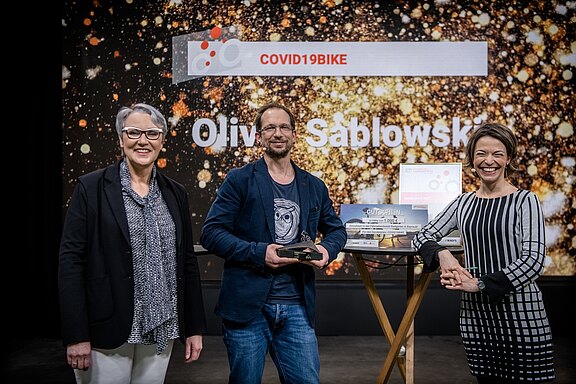 Drei Personen stehen auf einer Bühne. Ein Mann hält einen Preis und lächelt, während zwei Frauen neben ihm stehen. Im Hintergrund sind die Namen "COVID19BIKE" und "Olivier Sablowski" zu sehen. Auf einem Tisch liegen Gutscheine.