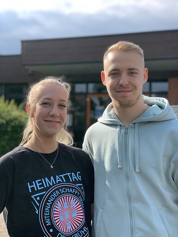 Zwei junge Menschen stehen draußen vor einem Gebäude. Die Frau trägt ein schwarzes T-Shirt mit dem Aufdruck "HEIMATTAG", während der Mann einen hellblauen Hoodie trägt. Beide lächeln freundlich in die Kamera.