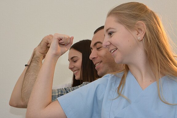 Drei Personen stehen nebeneinander und zeigen eine positive Körpersprache. Sie lächeln, während sie ihre geballte Faust in die Höhe strecken, was Freude und Selbstbewusstsein ausdrückt. Die Person ganz rechts trägt ein hellblaues Oberteil, während die anderen leger gekleidet sind.
