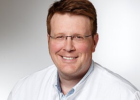 Porträt eines lächelnden Mannes mit Brille in weißem Kittel vor neutralem Hintergrund; Kopf-und-Schultern-Aufnahme, direkte Blickrichtung.