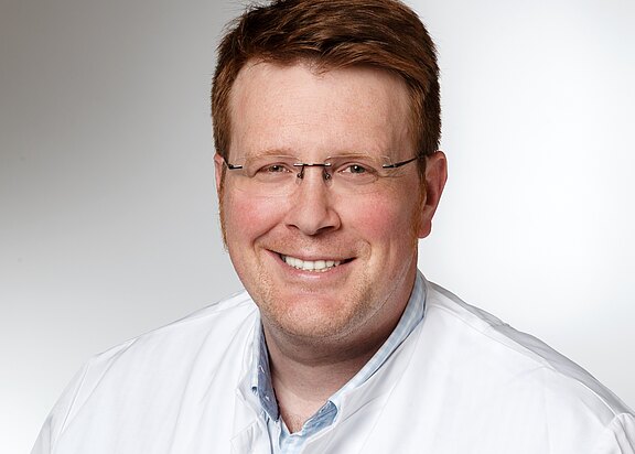 Porträt eines lächelnden Mannes mit Brille in weißem Kittel vor neutralem Hintergrund; Kopf-und-Schultern-Aufnahme, direkte Blickrichtung.