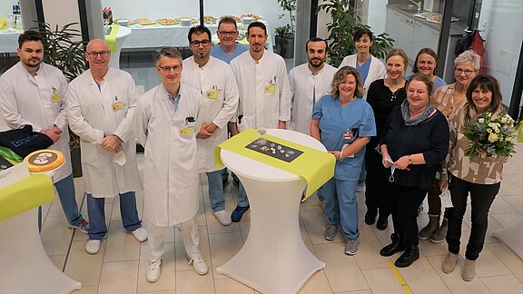 Eine Gruppe von zwölf Personen steht in einem modernen Raum zusammen, darunter Ärzte und Pflegekräfte. Sie lächeln und posieren um einen runden Tisch, auf dem dekorative Objekte und eine Torte platziert sind. Im Hintergrund sind Snacks sichtbar.