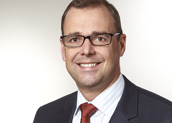 Porträt eines lächelnden Mannes mittleren Alters mit Kurzhaar und Brille. Er trägt dunklen Anzug, weißes Hemd und rote Krawatte. Neutraler, heller Hintergrund. Fokus auf das freundliche, professionelle Erscheinungsbild.