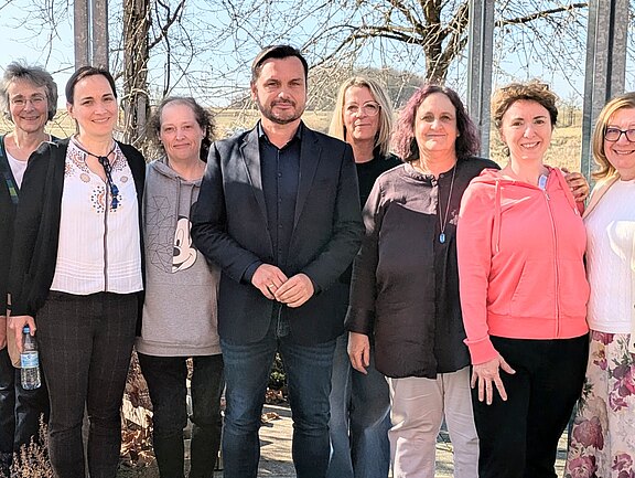 Gruppenfoto: Mehrere Erwachsene stehen nebeneinander und lächeln in die Kamera. Sie posieren im Freien bei sonnigem Wetter, vor kahlen Bäumen. Schwerpunkt: freundliche, gemeinschaftliche Stimmung.