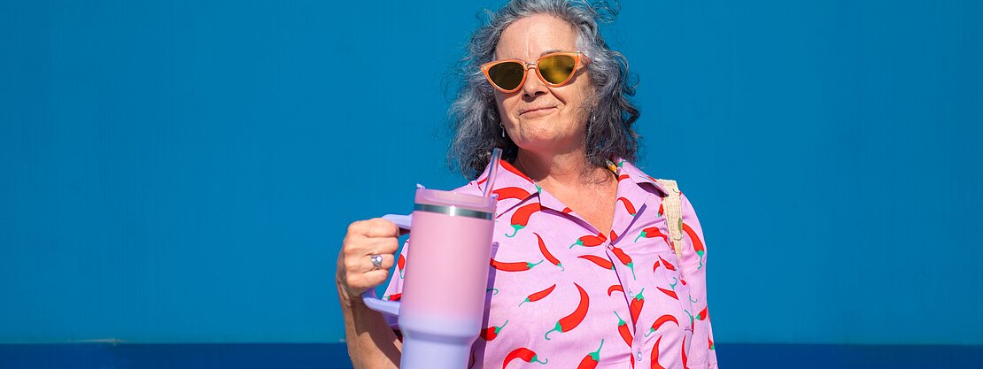 Ältere Frau mit orangefarbener Sonnenbrille vor knallblauer Wand. Sie trägt ein pinkes Hemd mit Chili-Muster und hält einen großen pastellfarbenen Trinkbecher mit Henkel. Ausdruck selbstbewusst, sommerliche Szene.