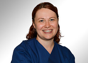 Dr. med. Daniela Opel Porträt einer lächelnden Frau mit rötlichen Haaren und Zahnspange, die blaue Pflege- oder OP-Kleidung trägt, vor hellem neutralem Hintergrund.