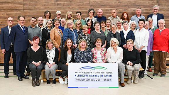 Gruppenfoto von etwa 40 Erwachsenen in einem Innenraum. Vorne ein Schild mit der Aufschrift: Klinikum Bayreuth GmbH – Medizincampus Oberfranken (Kliniken Bayreuth, Klinik Hohe Warte). Die Personen posieren lächelnd, teils sitzend, teils stehend, für ein offizielles Teamfoto.