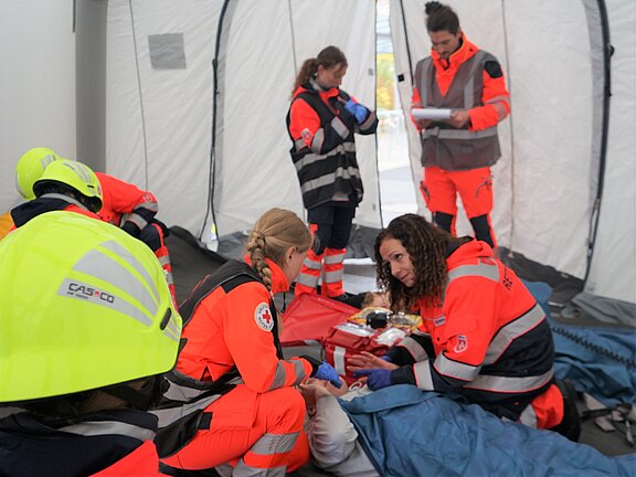 In einem Zelt arbeiten mehrere Rettungskräfte in leuchtend orangefarbenen Uniformen. Eine Person kümmert sich um eine verletzte Person, während andere aktiv Unterstützung leisten. Es herrscht eine konzentrierte, kooperative Atmosphäre, die auf Notfallhilfe hinweist.