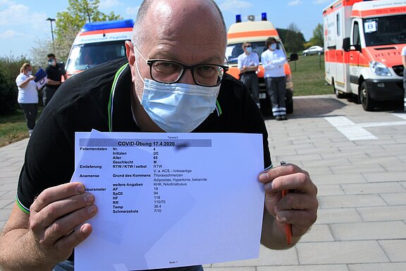 Ein Mann hält ein Dokument mit Patientendaten zur COVID-Übung vom 17. April 2020 in der Hand. Das Dokument enthält Informationen wie Name, Alter, Geschlecht und gesundheitliche Angaben. Im Hintergrund sind Rettungsfahrzeuge und weitere Personen im medizinischen Bereich zu sehen.