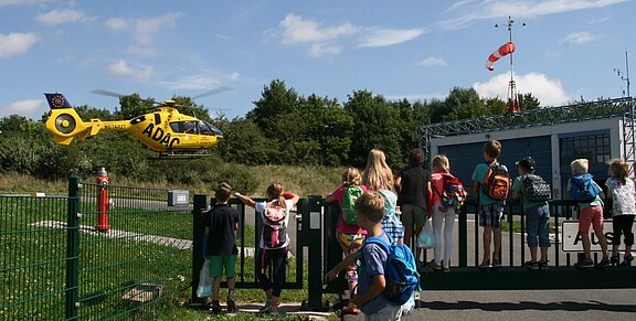 Eine Gruppe Kinder steht hinter einem Zaun und beobachtet einen gelben ADAC-Rettungshubschrauber, der nahe der Landefläche startet oder landet; ein Windsack zeigt die Windrichtung.