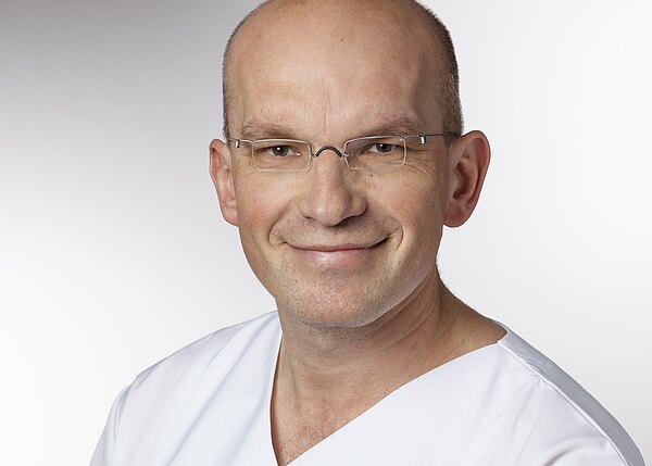 Porträt eines lächelnden, glatzköpfigen Mannes mit Brille in weißer Arbeitskleidung vor neutral hellem Hintergrund; Kopf und Schultern sind sichtbar.
