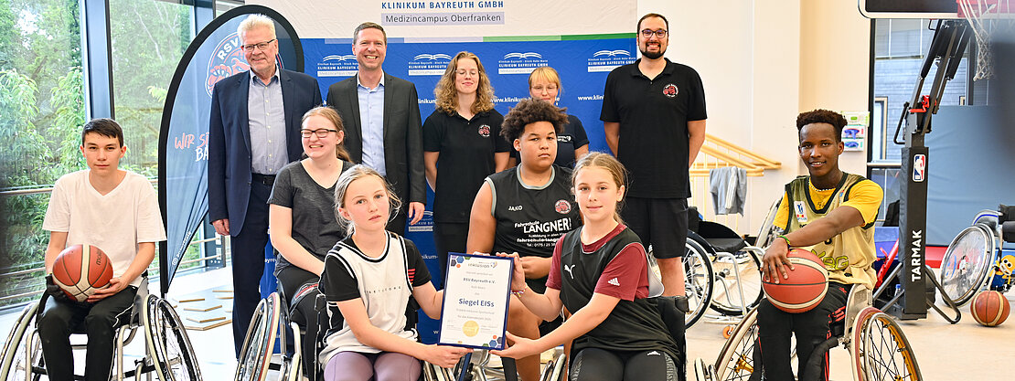 Die Abbildung zeigt eine Gruppe von Jugendlichen im Rollstuhl, die in einer Sporthalle zusammen sind. Einige halten Basketballs, andere präsentieren ein Zertifikat. Im Hintergrund stehen Erwachsene, die die Veranstaltung unterstützen. Die Atmosphäre vermittelt Gemeinschaft und sportlichen Geist.