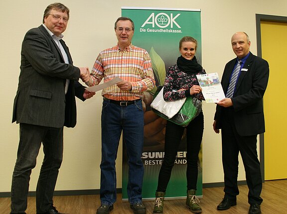 Vier Personen bei einer Urkundenübergabe: Links und rechts überreichen zwei Männer Dokumente. In der Mitte nehmen ein Mann und eine Frau Zertifikat und Geschenke entgegen. Im Hintergrund steht ein AOK-Banner.