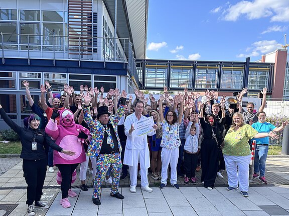 Große Gruppe von Menschen steht vor einem modernen Gebäude, lächelt in die Kamera und hebt die Arme. Viele tragen bunte Kostüme oder auffällige Outfits, einige Alltagskleidung. Die Szene wirkt wie eine fröhliche Gruppenaufnahme im Freien bei sonnigem Wetter.