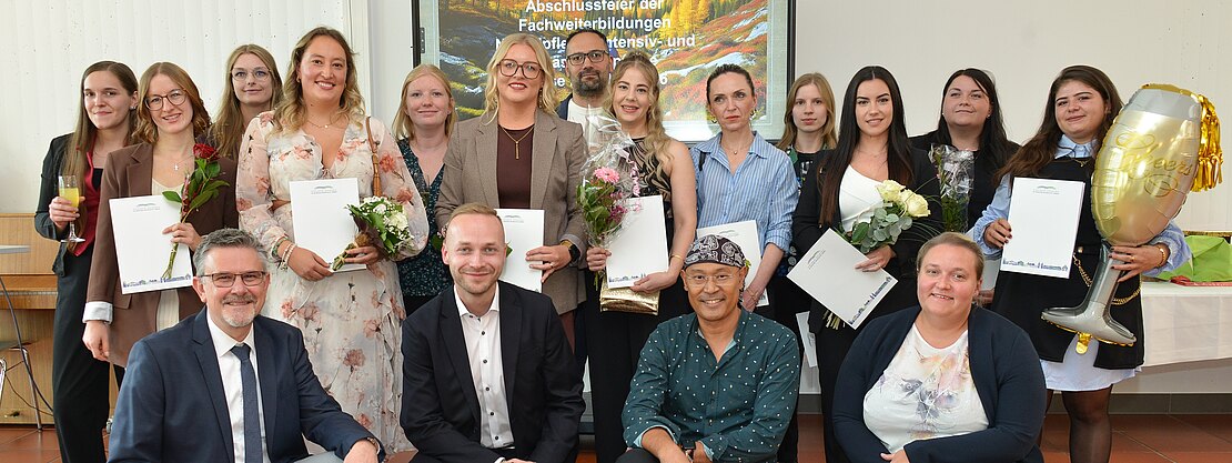 Gruppenfoto bei einer Abschlussfeier: Erwachsene posieren lächelnd in einem Saal, einige halten Urkunden und Blumen. Vorne sitzen mehrere Personen, dahinter steht eine Reihe. Feierlicher Anlass.