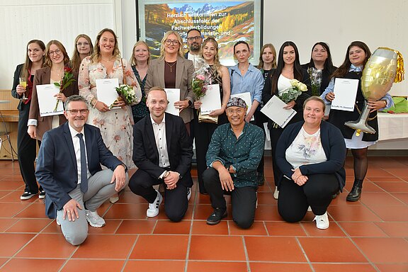 Gruppenfoto bei einer Abschlussfeier: Erwachsene posieren lächelnd in einem Saal, einige halten Urkunden und Blumen. Vorne sitzen mehrere Personen, dahinter steht eine Reihe. Feierlicher Anlass.