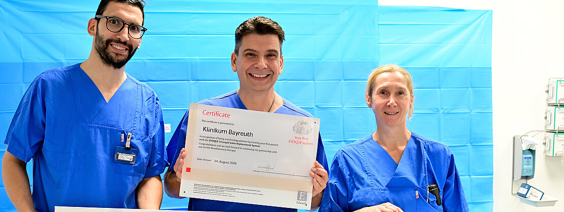Drei medizinische Fachkräfte in blauen OP-Kitteln posieren im Behandlungsraum. Die mittlere Person hält ein Zertifikat mit der Aufschrift Klinikum Bayreuth. Vor ihnen liegt eine lange Verpackung mit Evoque Transcatheter Delivery System – Präsentation eines neuen Medizinprodukts, feierliche Stimmung.