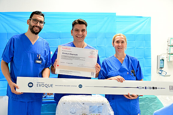 Drei medizinische Fachkräfte in blauen OP-Kitteln posieren im Behandlungsraum. Die mittlere Person hält ein Zertifikat mit der Aufschrift Klinikum Bayreuth. Vor ihnen liegt eine lange Verpackung mit Evoque Transcatheter Delivery System – Präsentation eines neuen Medizinprodukts, feierliche Stimmung.