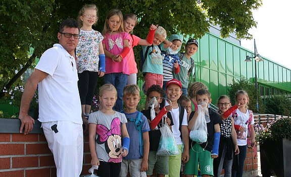 Gruppe fröhlicher Grundschulkinder steht mit einem Betreuer im Freien. Sie posieren auf einer Mauer neben Bäumen und einem modernen Gebäude; einige halten durchsichtige Beutel mit gesammelten Sachen. Ausflug oder Schulaktion, sommerliches Wetter.