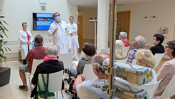 Eine Gruppe älterer Menschen sitzt in einem Raum und hört einem Arzt zu, der eine Präsentation hält. Neben ihm steht eine Assistenz, während ein weiterer Arzt im Hintergrund wartet. Auf einem Bildschirm wird "Vielen Dank für die Aufmerksamkeit!" angezeigt. Der Raum ist hell und freundlich gestaltet.