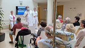 Eine Gruppe älterer Menschen sitzt in einem Raum und hört einem Arzt zu, der eine Präsentation hält. Neben ihm steht eine Assistenz, während ein weiterer Arzt im Hintergrund wartet. Auf einem Bildschirm wird "Vielen Dank für die Aufmerksamkeit!" angezeigt. Der Raum ist hell und freundlich gestaltet.
