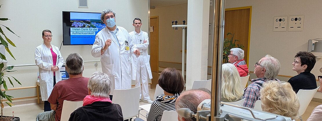 Eine Gruppe älterer Menschen sitzt in einem Raum und hört einem Arzt zu, der eine Präsentation hält. Neben ihm steht eine Assistenz, während ein weiterer Arzt im Hintergrund wartet. Auf einem Bildschirm wird "Vielen Dank für die Aufmerksamkeit!" angezeigt. Der Raum ist hell und freundlich gestaltet.