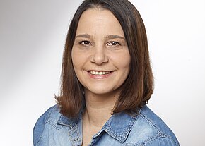 Porträt einer lächelnden erwachsenen Person mit schulterlangem braunem Haar vor neutralem Hintergrund, trägt ein blaues Jeanshemd.